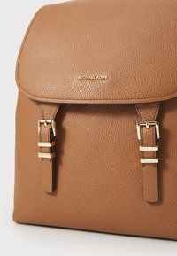 Sac à dos en cuir marron à texture granuleuse avec une fermeture à rabat, doté de quincaillerie dorée et de deux bretelles réglables. Logo Michael Kors en relief sur le devant.