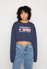Sudadera recortada de color azul marino con mangas largas, que presenta un logotipo blanco de "TOMMY" y un gráfico de bandera rectangular. Combinada con jeans de color azul claro.
