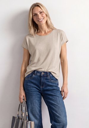 Uśmiechnięta kobieta z blond włosami ubrana w beżowy t-shirt i niebieskie dżinsy, trzyma paskowaną torbę na tle jasnej ściany.
