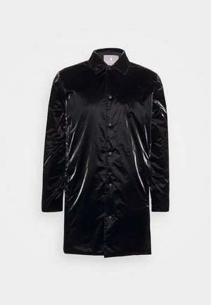 Long imperméable noir brillant à boutons, manches longues et col pointu, présenté sur un fond clair uni.
