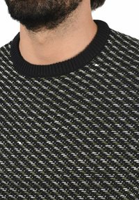 Suéter negro de punto con un patrón texturizado en verde y blanco, con un cuello negro acanalado, que muestra un diseño tejido.