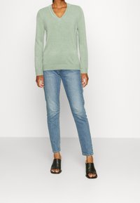 Maglione verde chiaro a maniche lunghe e scollatura a V, realizzato in materiale morbido, abbinato a jeans slim fit di lavaggio medio e mule nere con le punte scoperte.