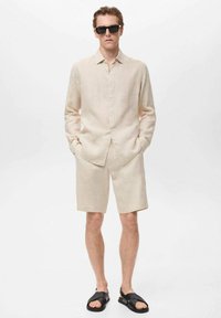 Camisa de lino beige claro de manga larga, combinada con pantalones cortos a juego. Sandalias negras y gafas de sol complementan el atuendo. Diseño simple y minimalista.