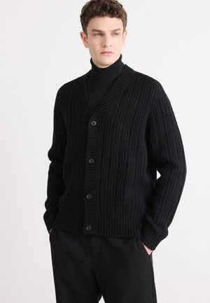 Cardigã preto canelado com decote em V, cinco botões na frente e mangas longas. Usado sobre um suéter de gola alta preto, combinado com calças escuras.