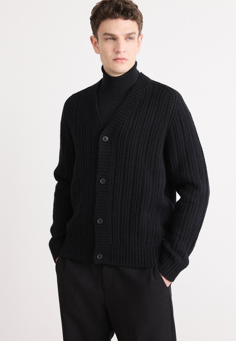 Μαύρο ριπτό καρδιάκι με V-λαιμόκοψη, πέντε κουμπιά μπροστά και μακριά μανίκια. Φοριέται πάνω από ένα μαύρο τσιμπούκι, συνδυασμένο με σκούρα παντελόνια.