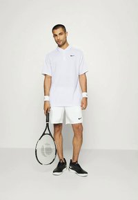 Hvit polo-shirt med svart Nike-logo, kombinert med hvite shorts og svarte joggesko. Holder en tenniskølle med mønstret netting.