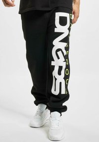 Schwarze Sweatpants mit großem weißen "DNGRS"-Schriftzug und gelber Akzentseite. Mit Bündchen an den Knöcheln und lockerer Passform, aus weichem Material gefertigt.