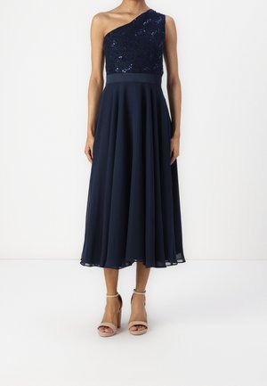 Femme portant une robe midi asymétrique bleu marine avec un haut à sequins et une jupe fluide, associée à des sandales à talons hauts couleur nude.
