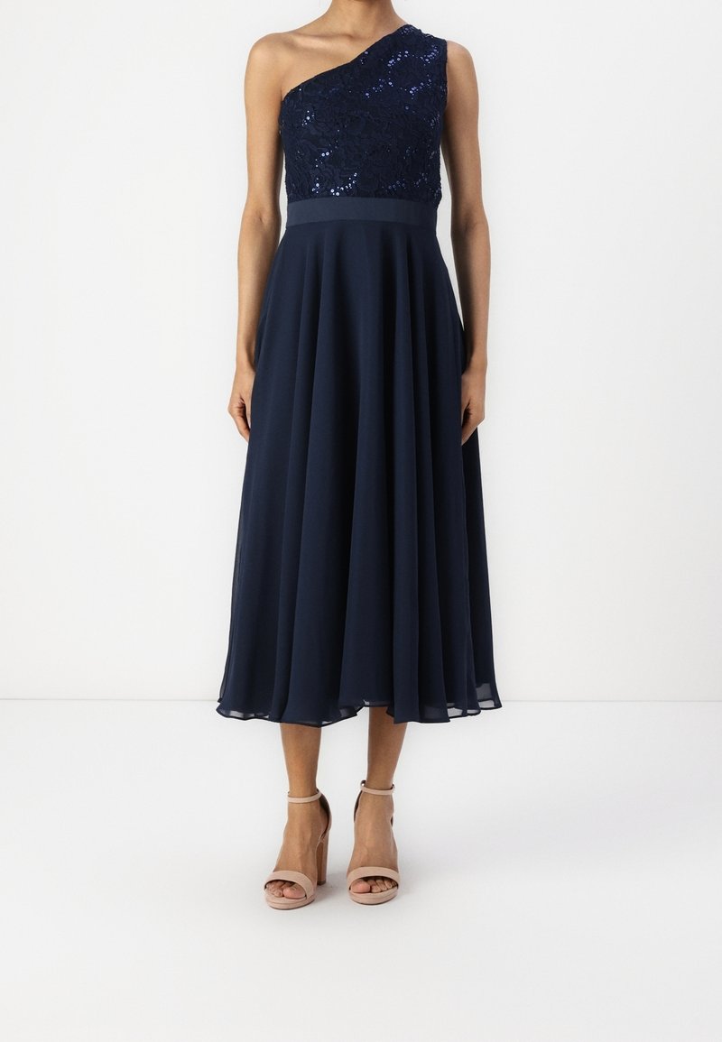 Femme portant une robe midi asymétrique bleu marine avec un haut à sequins et une jupe fluide, associée à des sandales à talons hauts couleur nude.