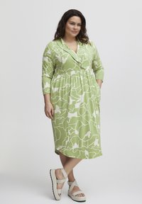 Vestido midi verde con un patrón abstracto en blanco, con mangas largas, cuello y bolsillos. Sandalias de color claro con suelas gruesas.