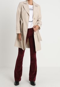 Beige trenchcoat med dubbelknäppt design, stora kragar och matchande bälte, bärs över en vit grafisk t-shirt och burgunderfärgade kordbyxor med utsvängda ben.
