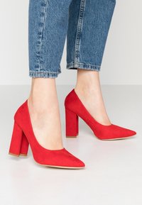 Rote Wildleder-Pumps mit spitzem Zeh und klobigem Absatz, die eine glatte Textur und ein elegantes Design präsentieren.