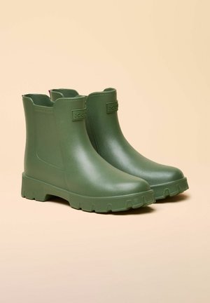 Paire de bottes de pluie en caoutchouc vert foncé pour cheville, avec semelles texturées et panneaux élastiques latéraux, posées sur un fond beige.