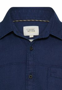 Męska granatowa koszula wykonana z fakturowanej tkaniny, wyposażona w kołnierzyk zapinany na guziki oraz jedną kieszeń na piersi. Wnętrze z widoczną metką marki.