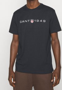 Camiseta de algodón azul marino con el logo en blanco "GANT 1949" y diseño de escudo en el pecho. Manga corta y cuello redondo.