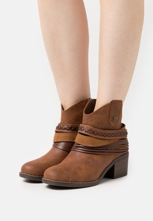 Jambes inférieures portant des bottines marron à talons carrés avec des brides tressées et enveloppées décoratives, sur fond blanc uni.