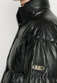 Czarna pikowana kurtka puchowa ze skóry z wysokim kołnierzem, pikowaną teksturą oraz złotą naszywką z logo na lewym rękawie. Przednie zapięcie na zatrzaski.