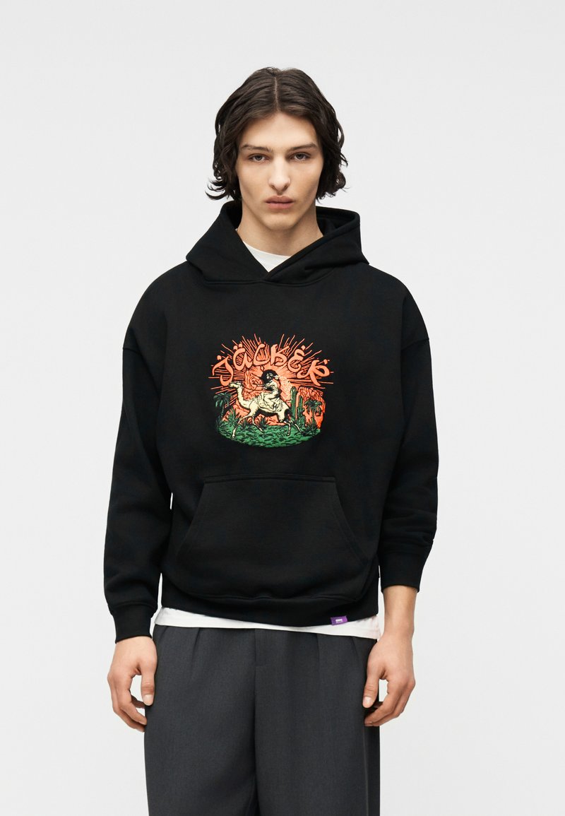 Νεαρό άτομο με μεσαίου μήκους σκούρα μαλλιά, φοράει μαύρη μπλούζα με κουκούλα με πολύχρωμο σχέδιο κάκτου και αλόγου, γκρι παντελόνι, στέκεται μπροστά σε απλό φόντο.