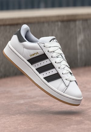 Biały sneaker z czarnymi paskami i napisem "CAMPUS", białe sznurowadła oraz gumowa podeszwa, przedstawiony w powietrzu na tle zewnętrznego otoczenia.