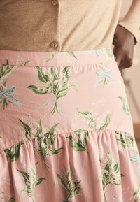 Cardigan camel cu nasturi, băgat într-o fustă roz cu talie înaltă, cu model verde de frunze și flori albe, purtat de o persoană cu piele închisă la culoare.