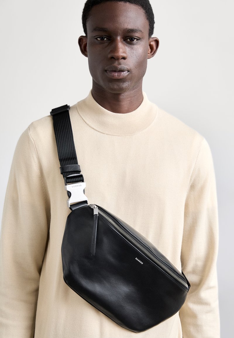 Sac en cuir noir en bandoulière avec une texture lisse, doté d'une fermeture éclair argentée et d'une sangle noire ajustable. Design minimaliste avec détail de logo.