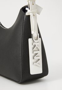 Borsa nera con texture e forma curva, dotata di una tracolla bianca e di un'etichetta rettangolare bianca con le lettere "DKNY" in rilievo argentato.