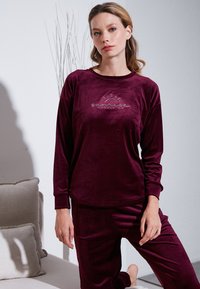 Burgundska velur lounge odjeća koja se sastoji od dugih rukava majice s grafikom sunca i odgovarajućih hlača, glatke teksture, opuštenog kroja i okruglog izreza.