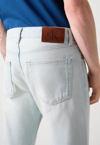 Jeans din denim de un albastru deschis, cu o croială slim, având un logo din piele și detalii de cusătură în contrast. Buzunarele din spate sunt vizibile.