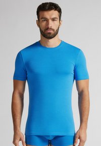 Kratší rukáv modré tričko vyrobené z měkké, elastické tkaniny. Design s kulatým výstřihem. Model má na sobě odpovídající modré kraťasy. Hladká textura bez viditelných vzorů.