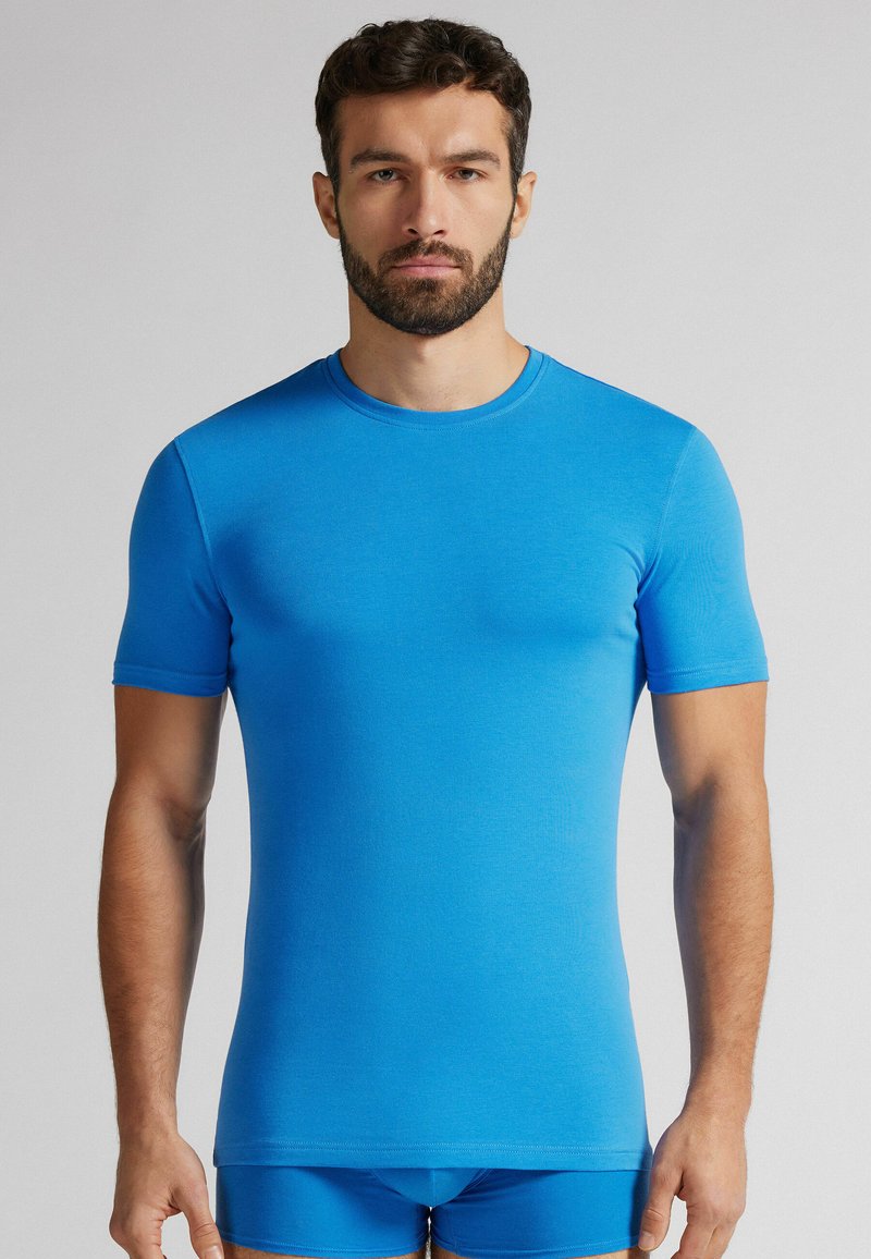 Kratší rukáv modré tričko vyrobené z měkké, elastické tkaniny. Design s kulatým výstřihem. Model má na sobě odpovídající modré kraťasy. Hladká textura bez viditelných vzorů.