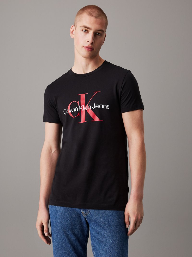 Giovane uomo con capelli corti che indossa una t-shirt nera Calvin Klein Jeans e jeans blu su uno sfondo grigio uniforme.