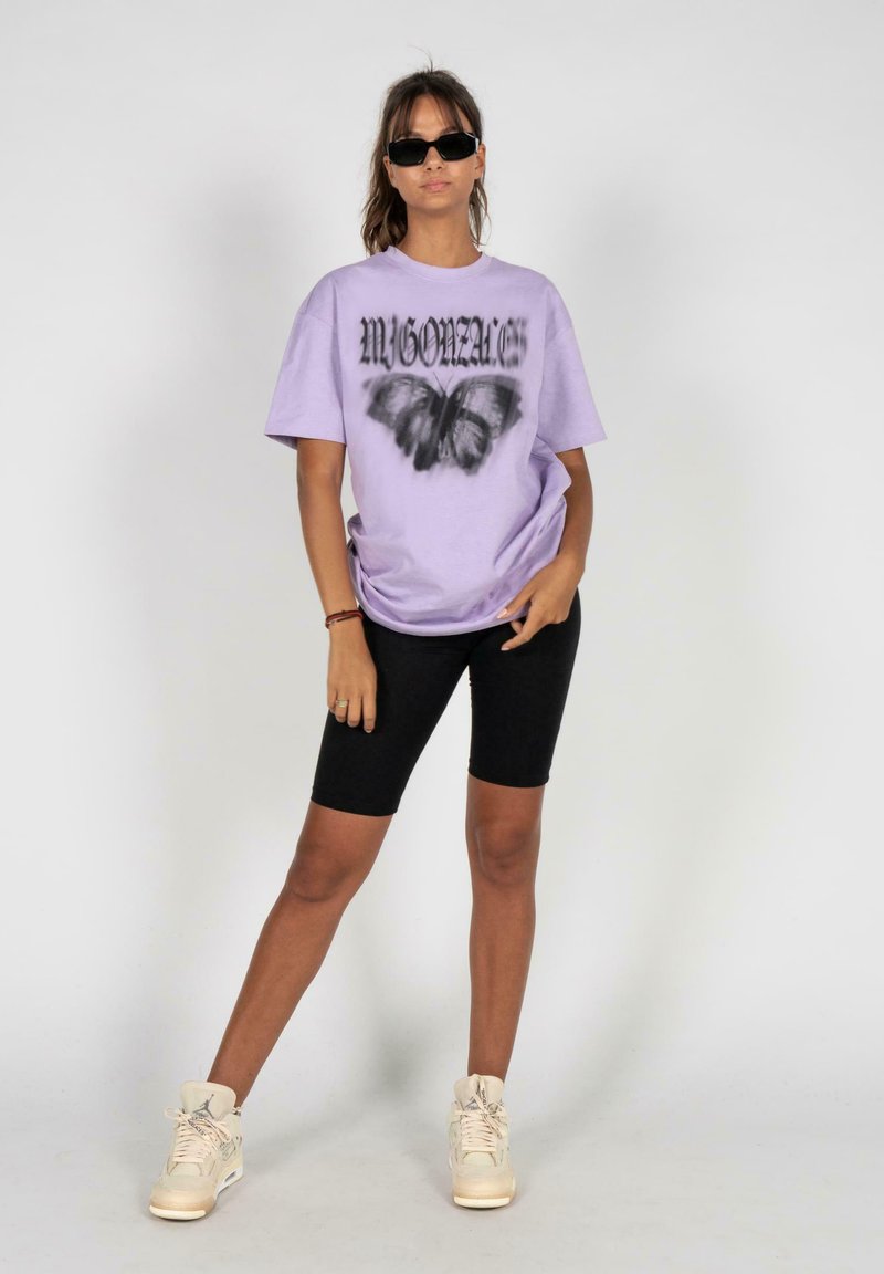 Lila übergroßes T-Shirt mit einem Schmetterlingsgrafik und schwarzem Text, kombiniert mit schwarzen Radlerhosen und beigen High-Top Sneakern.
