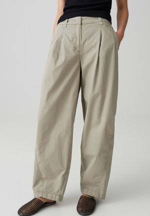 Femme portant un pantalon beige ample avec des plis et des poches, associé à des chaussures noires en mesh sans lacets et un top noir sans manches.