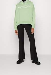 Sudadera con capucha en verde menta con texto de marca en blanco, combinada con pantalones acampanados negros y zapatos negros, sobre un fondo neutro.