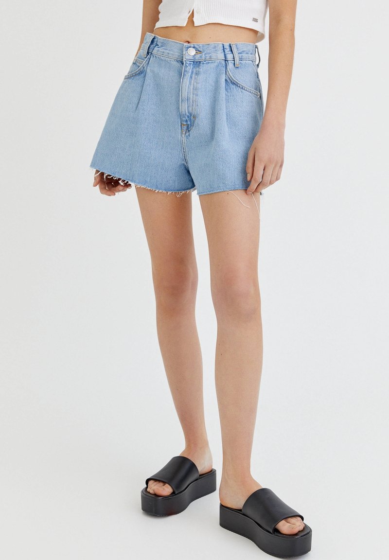 Femme portant un short en jean taille haute bleu clair avec ourlet effiloché, un top blanc court, et des sandales à plateforme noires, debout sur fond blanc.