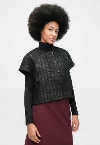 Mlada žena s afro frizurom nosi crni kratki kardigan teksture kratkih rukava preko crnog rolaka i burgundske suknje na bijeloj pozadini.