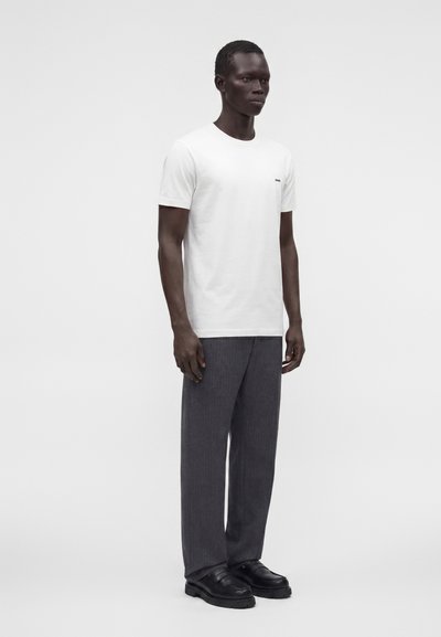 Modèle masculin debout portant un t-shirt blanc uni à manches courtes, un pantalon ample gris foncé et des chaussures en cuir noires sur fond clair.