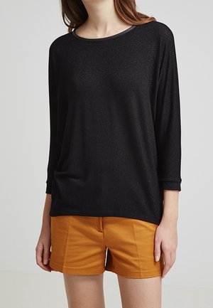 Woman wearing a black long-sleeve top and mustard yellow shorts, standing against a plain light background.