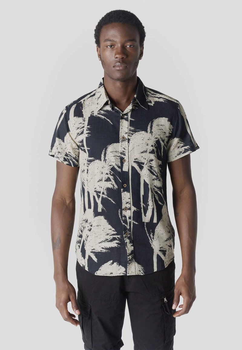 Homme portant une chemise noire à manches courtes avec un motif de palmiers beiges et un short cargo noir, debout devant un fond gris clair uni.