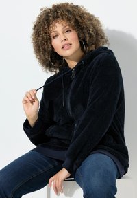 Kvinde med krøllet hår iført store hoop øreringe, en marineblå fuzzy zip-up hættetrøje og blå jeans, siddende og holder i hættetrøjens snor.