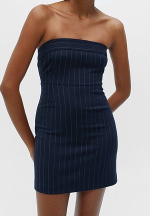 Kvinde iført en ærmeløs, stropløs mini-kjole i navyblå med tynde, lodrette hvide nålestriber, stående mod en ensfarvet hvid baggrund.