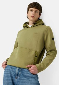 Olijfgroen hoodie met een voorzak, verstelbare trekkoord en zwart logo-opdruk op de mouw. Gepaard met blauwe spijkerbroek.