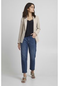 Blazer beige su un top nero con bordo in pizzo abbinato a jeans in denim blu, completato da sandali con tacco a blocco arricciati color verde salvia.