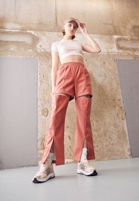 Byxor i mjukt, persikofärgat tyg med tryckknappsdetaill och sidofickor, parat med beige sneakers och vita strumpor, mot en neutral bakgrund.