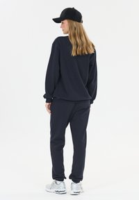 Vrouw met lang blond haar, gekleed in een zwarte pet, een zwart sweatshirt, zwarte joggers en witte sneakers, staande zijwaarts tegen een witte achtergrond.