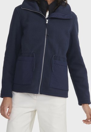 Kvinde iført en marineblå fleecejakke med lynlås og to forlommer samt hvide bukser, stående foran en ensfarvet baggrund.