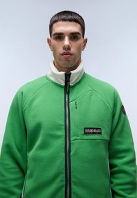Green fleece jacket with high collar, zip closure, and navy accents. Features a patch logo on the chest and textured fabric.