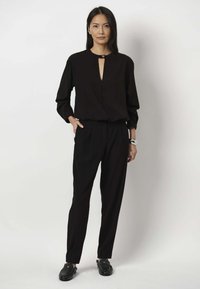 Schwarze Bluse mit einem Schlüssellochausschnitt, kombiniert mit schwarzen Hosen. Der Stoff ist glatt, mit langen Ärmeln und einer lässigen Passform. Schwarze Schuhe runden den Look ab.