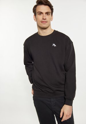 Ung mand stående mod en hvid baggrund iført en sort sweatshirt med "Mr." broderet på brystet og sorte bukser.