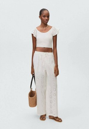 Top bianco a maniche voluminose, abbinato a pantaloni bianchi a gamba larga con ritagli floreali. Il modello tiene una borsa intrecciata e indossa sandali.